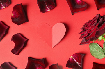 Passion concept for Valentine's day with dark red rose, petals and a paper heart at the centre of photo on red background.