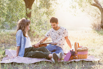 On a beautiful sunny day, a couple of young lovers, makes picnic on grass among olive groves in Tuscany, Italy. Man leans on guitar while talking with his girlfriend