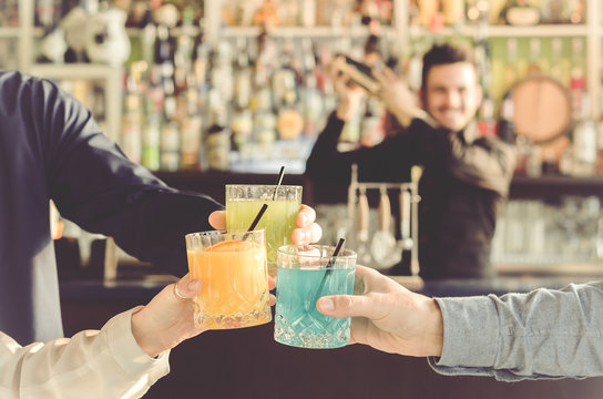 three friends are toasting with their drinks in hand during a party while the barman is shaking - people, drinks, party and lifestyle concept