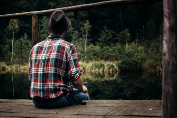 stylish hipster traveler sitting at sunny forest and lake in the