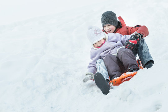 Sibling brother and little sister enjoying fast ride down snowy hill on orange snow slider