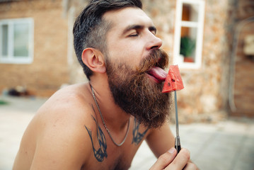 bearded man without clothes with a big juicy ripe watermelon in hands on a background of flowering garden illuminated by bright sunshine