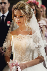 Naklejka premium Beautiful emotional blonde bride holding candle in church in whi