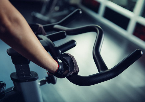 Close-up of young sportswoman hands in gloves working out on bicycle indoors in the gym. Fitness, training and lifestyle concept.