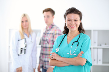 Beautiful young smiling female doctor standing at hospital with doctor and patient on the background. Medical concept.