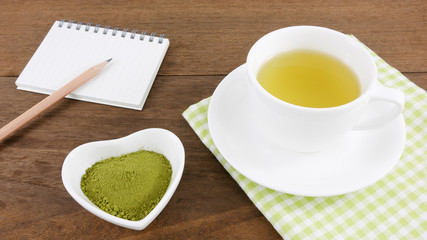 Obraz na płótnie Canvas The Japanese matcha green tea powder on ceramic heart shaped bowl and cup of hot green tea and note book with pencil on wooden planks.