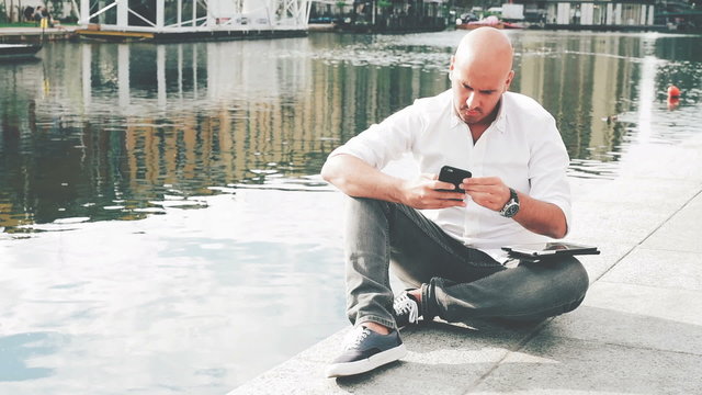 Young bald handsome caucasian businessman sitting on a river shore using a tablet on his knee tapping touchscreen then switching to smartphone handhold – technology, business, communication concept