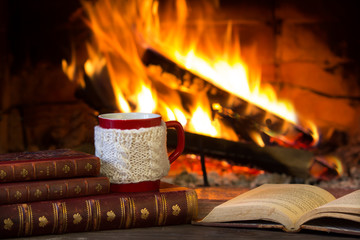 Cup of hot drink and antique books in front of warm fireplace. Red mug in white knitted mitten standing near fire. Magical relazed cozi atmosphere.