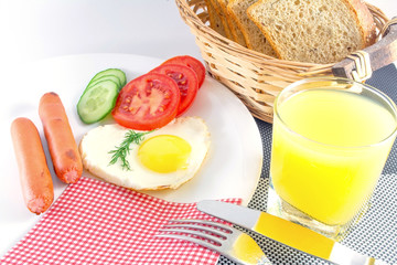 breakfast on a white plate on a black and white table Fried egg in a heart, fried sausage, fresh vegetables chopped cucumbers and tomatoes, juice, sliced bread, knife and fork on either side, napkin