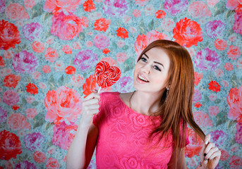 Fototapeta premium Studio portrait of beautiful smiling young woman in a pink dress holding heart shape red lollipop in front of colorful background