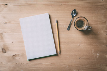 A recycled paper notebook checked with a black pencil with the eraser at the top and a cup of coffee with a spoon, are arranged on a wooden table. View from the top