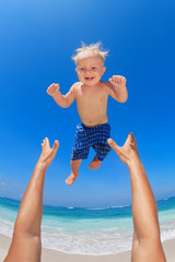 Family swimming fun on white sand sea beach and blue sky - father hands tossing up baby boy into mid air and catching. Child outdoor activity, active lifestyle on summer vacation in tropical island.
