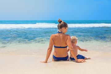 Happy family - mother and small baby son sit on sand beach and look at sea surf before swimming in clear water. Active parents and people outdoor activity on tropical summer vacations with child.