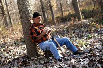 Portrait of senior lumberjack in forest sitting on a tree stump. He is resting and having lunch break.