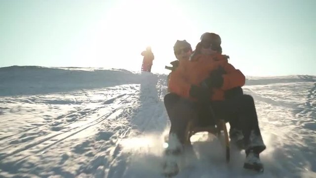 4k footage, happy best ager couple tobogganing together on one sled on sunny winter day while friends waiting on snowy hill
