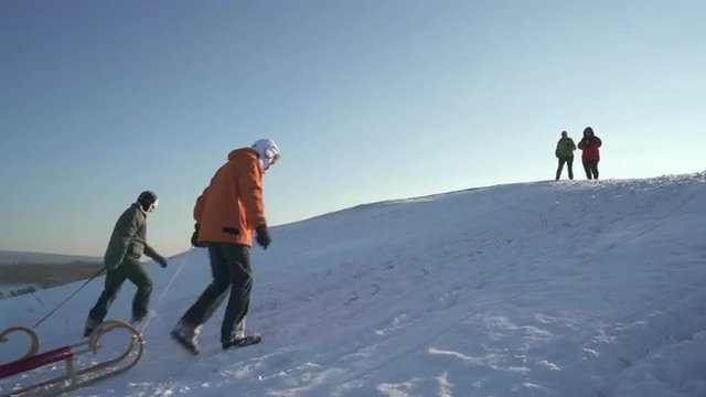 4k footage, two best ager men in winter clothes on sunny vacation day walking up snowy hill with sledges their wives awaiting them 
