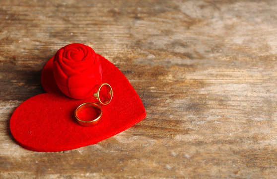 Red velvet silk rose box and weddings rings with heart on wooden background closeup