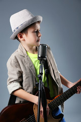 Little boy playing guitar and singing with microphone on grey background