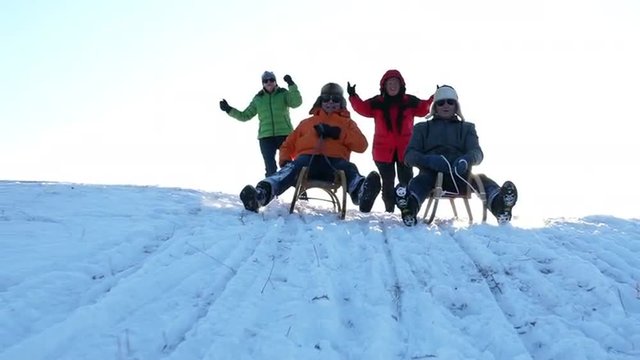 slow motion happy best ager couples enjoying sunny winter day outdoors on snowy hill, wives pushing and cheering their tobogganing laughing husbands
