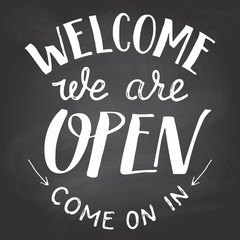 Obraz premium Welcome we are open. A welcome sign for cafes or shop visitors on blackboard background with chalk. Hand lettering