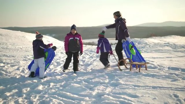 4k footage, mother with three young daughters walking outdoors in snow with bobsleds and sled up snowy hill
