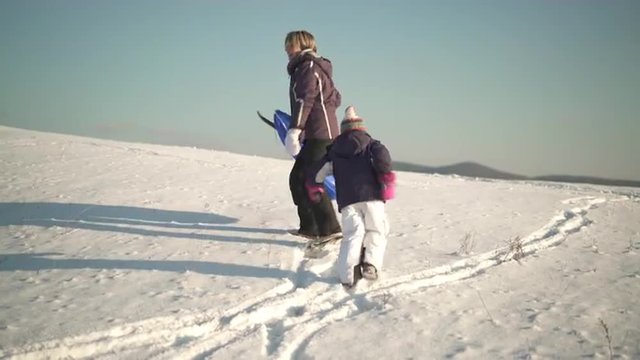 4k footage, mother and daugter walking up snowy hill with bobsled on sunny winter day
