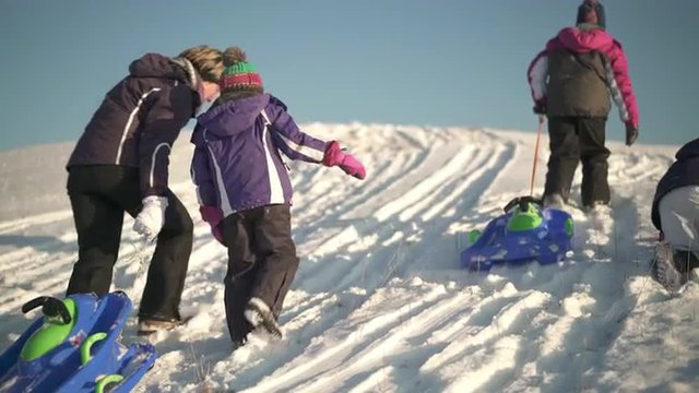 4k footage, mother walking up snowy hill with her three daughters and bobsleds on sunny winter day
