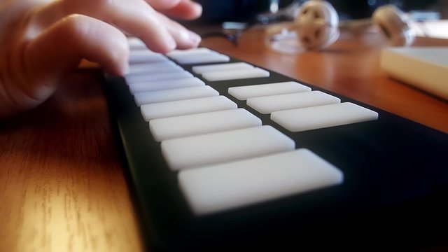 Hand fingers playing on a MIDI keyboard with light up buttons, headphones and notebook laying aside defocused
