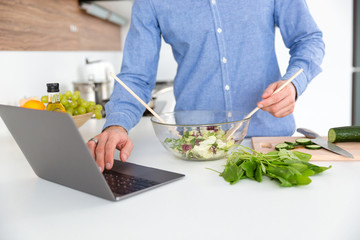 Man using laptop and making salad in glass bowl