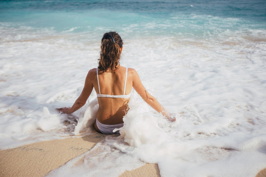 Back view of young woman in swimsuit sitting at white sand beach