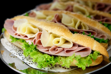 Yellow sliced cheese, ham and salad white  baguettes on doily and stainless steel tray. Black background.