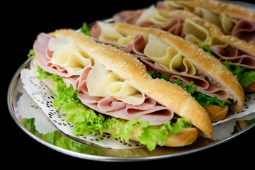 Yellow sliced cheese, ham and salad white  baguettes on doily and stainless steel tray. Black background.