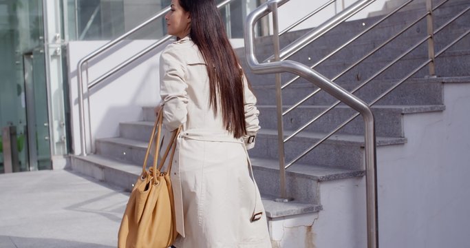 Elegant woman walking through town viewed from the rear in a white coat about to walk up a flight of outdoor stairs