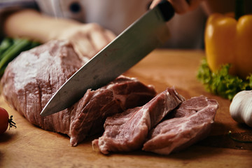 Male chef with knife cutting raw meat for steaks preparation. Piece of pork with vegetables and garlic on wooden table. 