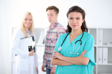 Fototapeta premium Beautiful young smiling female doctor standing at hospital with doctor and patient on the background. Medical concept.
