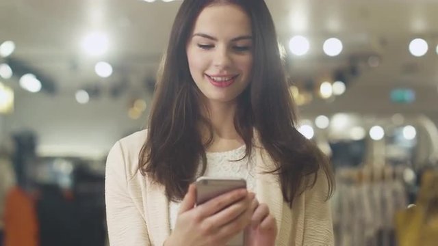 Happy young brunette woman is standing in a department store and using a smartphone. Shot on RED Cinema Camera.