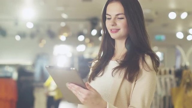 Happy young brunette girl is standing in a department store and using a tablet computer. Shot on RED Cinema Camera.