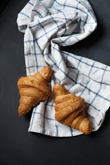 Fototapeta na wymiar Two fresh croissant lying on a blackboard. Also, there is twisted linen towel. Croissants are delicious and appetizing. Towel is white with blue pattern.