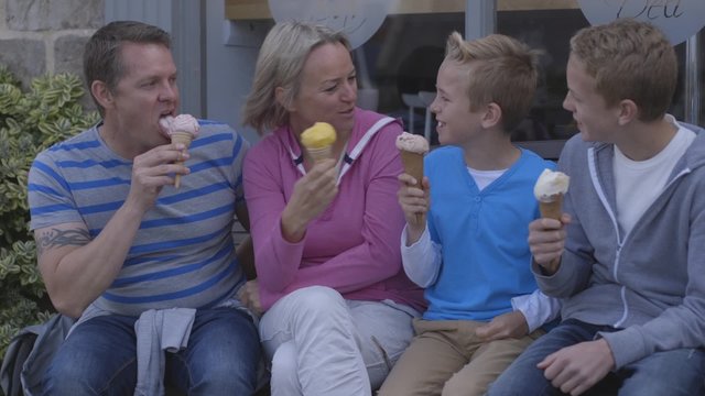 Family of four enjoying ice-cream sitting on a bench outside. They are wearing casual clothing and laughing.