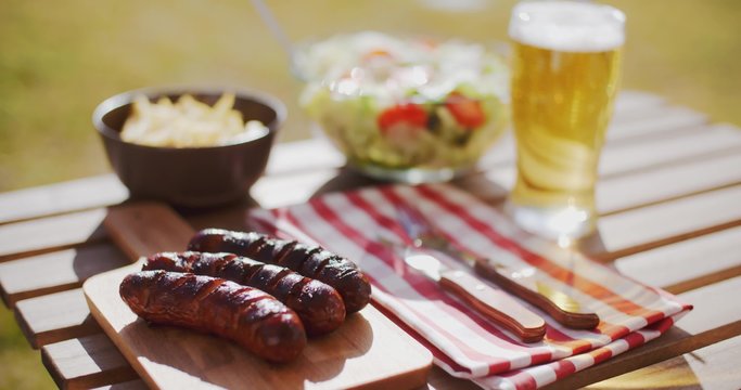 Grilled sausages and fresh green salads with a bowl of potato chips for a healthy summer picnic on an outdoor wooden table served with a beer.