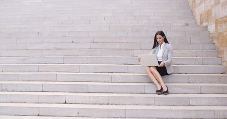 Fototapeta na wymiar Pretty young female worker with brown hair sitting alone on marble staircase with laptop outdoors