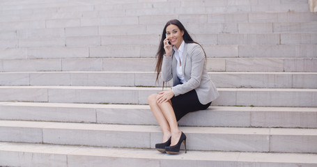 Fototapeta na wymiar Happy beautiful female executive sitting alone on outdoor marble stairs talking on her phone