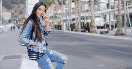 Trendy attractive young woman in a denim outfit perching on a wall on an urban promenade talking on her mobile and smiling at the camera