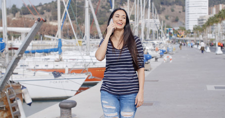 Attractive fashionable young woman on her mobile phone walking along a waterfront promenade at a marina with moored boats
