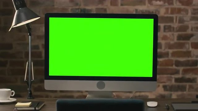 Footage of a computer monitor with green screen standing on a table next to a tablet, lamp, coffee cup, notebook and mouse in a loft. Shot on RED Cinema Camera.