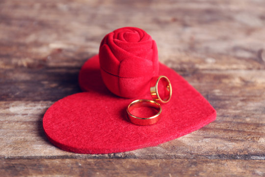 Red velvet silk rose box and weddings rings with heart on wooden background closeup