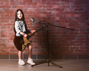 Beautiful little girl with microphone and guitar on brick wall background