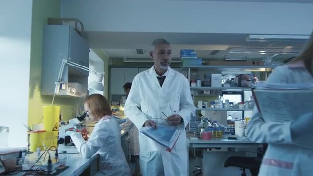 Senior scientist is walking with documents in a laboratory where colleagues are working.  Shot on RED Cinema Camera.