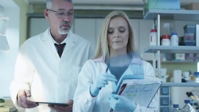 Senior and female scientists with papers and tablet are walking and having a conversation in a laboratory.  Shot on RED Cinema Camera.