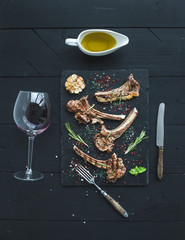 Grilled lamb chops. Rack of Lamb with garlic, rosemary, spices on slate tray, wine glass, oil in a saucer over black wood background